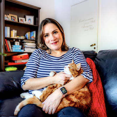 En mörkhårig kvinna sitter i en soffa med en katt i famnen. 