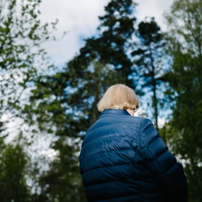 Äldre i blå jacka utomhus omgiven av trädkronor. Sirkka Kurkela i Esbo i maj 2020.