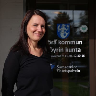Kvinna står framför en dörr med ett kommunvapen. Hon ler och tittar in i kameran.
