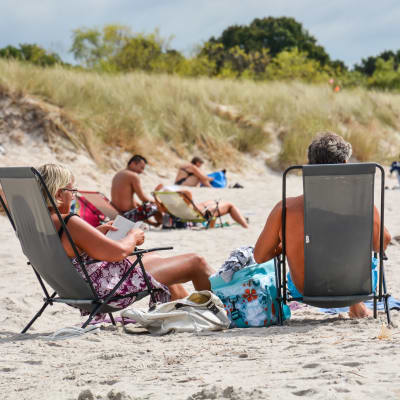 Ett medelålders par solar i varsin solstol på en strand. I bakgrunden flera solbadare.