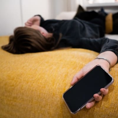 Anonyymi nainen makaa vuoteellaan puhelin kädessään.