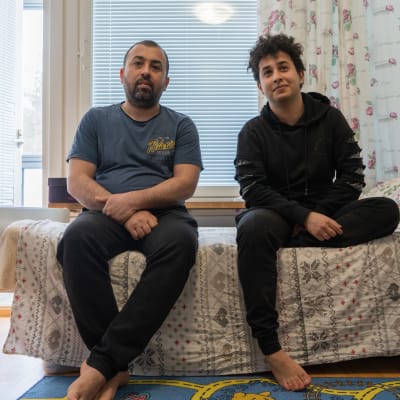 Qosai sitter med sonen Zein på en säng framförett fönster med persienner och blommiga gardiner.