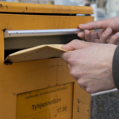 Kädet sujauttamassa ruskeaa kirjekuorta postin oranssiin laatikkoon.