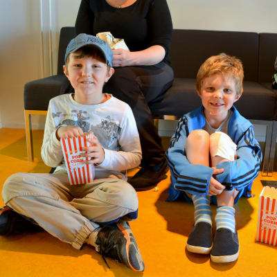 Två pojkar sitter på golvet framför en bänk och äter popcorn