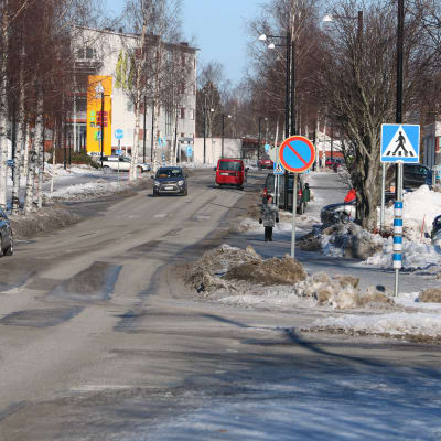Stadscentra. Tre bilar kör på en väg och på trottoaren går några människor. Det är snö på marken. I förgrunden syns också ett flertal trafikmärken.