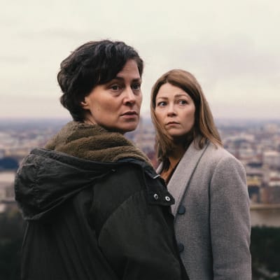 Rita (Lotta Lehtikari) ja Laura (Niina Koponen) katsovat olkansa yli huolestuneen näköisinä kaupunkimaisena taustallaan.