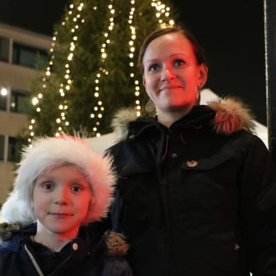 Barn med tomtemössa står med sin mor framför julgran som är upplyst.