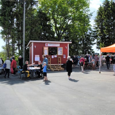 Bilden föreställer ett sommartog på Vessö. På bilden syns ett cafè, samt människor som besöker torget.