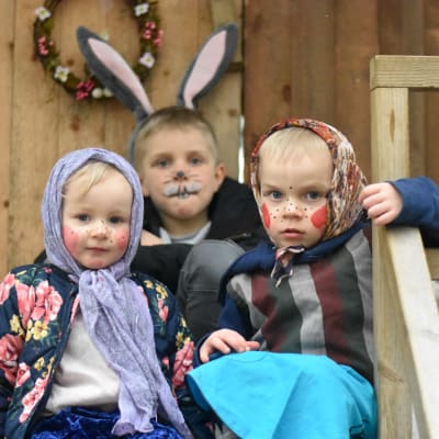 Tre små barn sitter på trappan till en träkoja. En är utklädd till hare och de två andra till häxor.