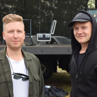 Två unga män står framför en konsertscen. Utomhus.