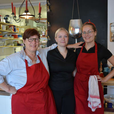 Tre kvinnor står i en restaurang, två har röda förkläden.