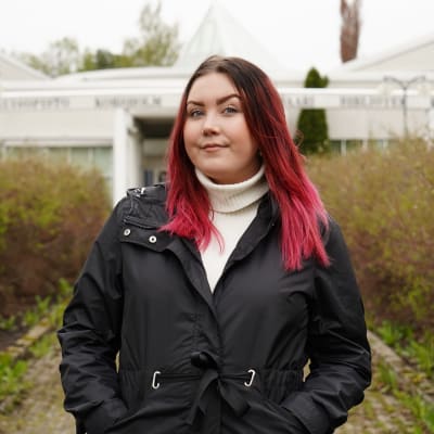 Ung kvinna med rött hår ler in i kameran. I bakgrunden syns Korsholms kulturhus.