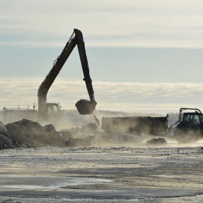 en grävskopa lassar äver muddringsmassor i ett traktorsläp på isen. 