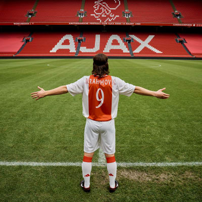 En fotbollsspelare fotad bakifrån står med armarna utsträckta framför en läktare där det står AJAX.