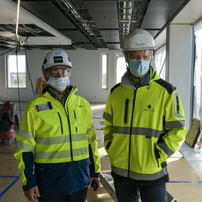 Två män i arbetskläder, hjälm, skyddsglasögon och munskydd står i ett stort rum som är under konstruktion.