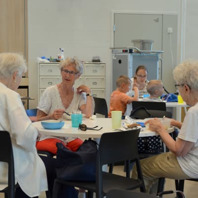 Tre äldre damer i vita skjortor sitter runt ett bord och äter och pratar 