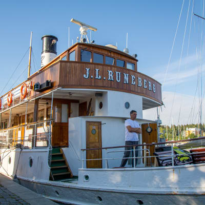 Båt som det står "J. L Runeberg" på förtöjd vid en hamn. En person står på däcket. 