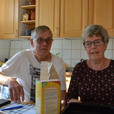 John och Birgitta Åsvik sitter i sitt kök. Birgitta bär en mörk blus och john en vit t-skjorta. 