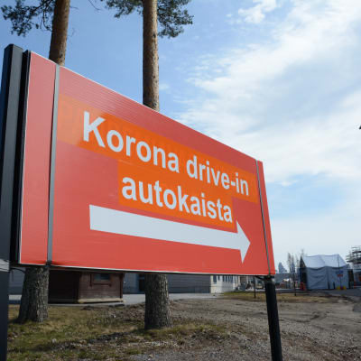 Iso oranssi kyltti Koronatestauksesta Kajaanin keskussairaalan pihassa. Taka-alalla näkyy teltta, jossa drive-in -testaukset suoritetaan.