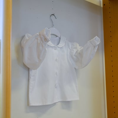 En vit kragskjorta för barn hänger i en bokhylla på en klädhängare. Skjortans ärmar är utsträckta. 