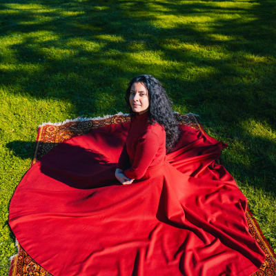 Nainen punaisissa vaatteissa istuu matolla ulkona puistossa.