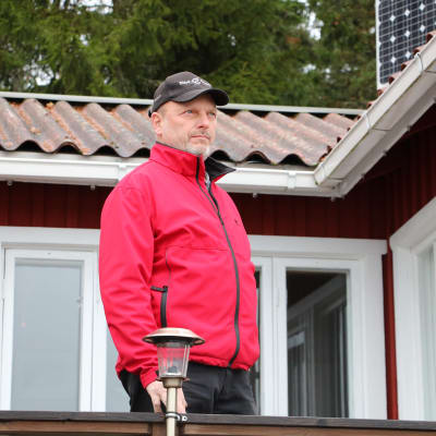 En medelålders man i röd jacka och med keps på huvudet står vid ett terrassräcke vid en röd sommarstuga.