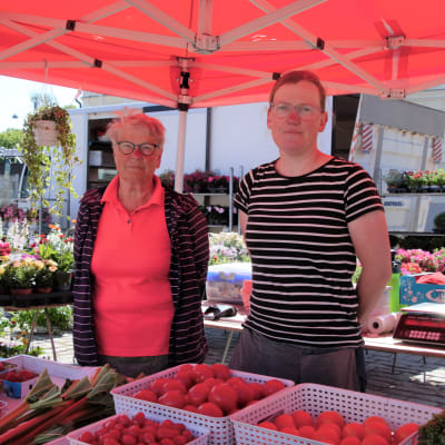 På bilden står Gunda Lindholm och Eva Nylund. Gunda har en röd skjorta på sig och glasögon. Eva har en vit-svart randig skjorta på sig. I bakgrunden ser man blommorna från deras blomsterhandel.