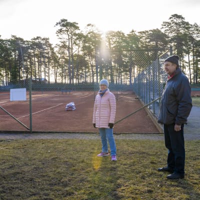 En kvinna och en man står bredvid tennisbanor. Det är ute, tidig vår och gäller utomhusbanor.