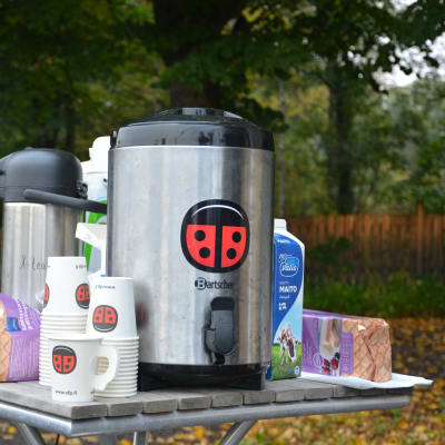 En stor kaffetermos och engångsmuggar med SFP-logo, mjölk och småbråd på ett litet trädgårdsbord, höstlöv i bakgrunden.