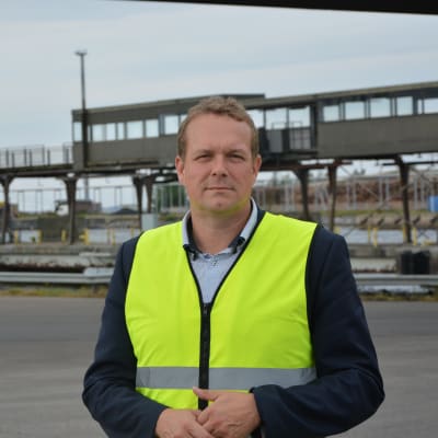 Matti Esko är vd för Kvarken ports. På bilden bär han en gul skyddsväst och står där fordon kör ombord på Wasaline. I bakgrunden ser man en gammal passagerargång, en pir och ett fraktfartyg.