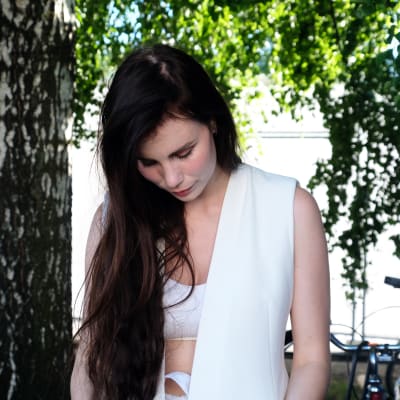 Den svenska artisten Skott poserar bakom scenen på årets Flow-festival (2017), hon är klädd helt i vitt och har långt, mörkt hår. Hennes blick är riktad mot marken.