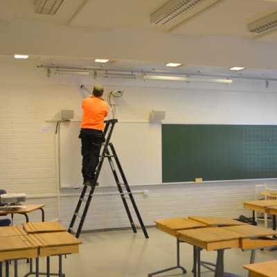 En man i orange tröja står på en stege och lägger tätning vid taklisten i ett klassrum.