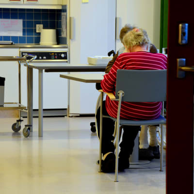 Ett par äldre personer sitter vid ett bord. I bakgrunden syns ett kök.