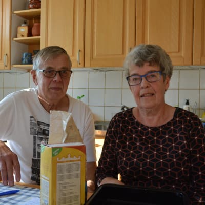 John och Birgitta Åsvik sitter i sitt kök. Birgitta bär en mörk blus och john en vit t-skjorta. 