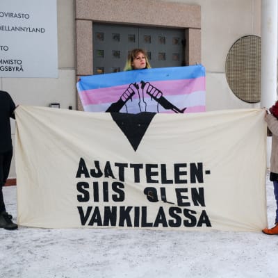 Tre personer håller i en vit banderoll med texten "Ajattelen - siis olen vankilassa".