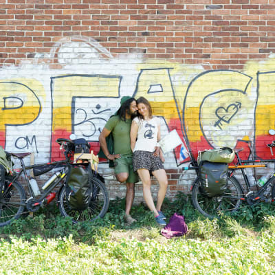 En man och en kvinna står framför en grafittimålning som föreställer ordet "peace". Bredvid sig har de sina cyklar som är fullpackade med väskor. 