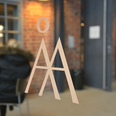 Åbo Akademis textlogo, ett Å och ett A klistrat på en glasdörr, insidan av huset med röd tegelvägg i bakgrunden.