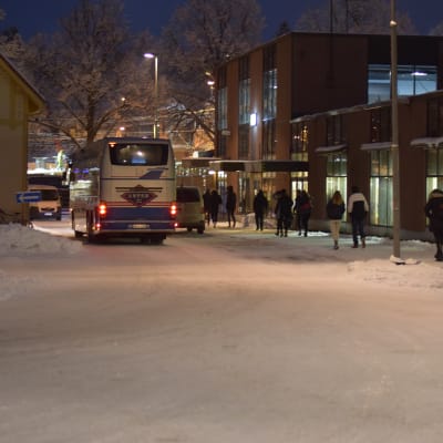 En buss kör iväg från busstation. Man ser bara baklyktorna. Vinterlandskap. Några ungdomar som stigit av bussen går på trottoaren.