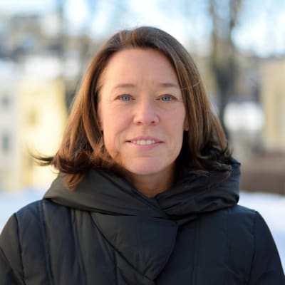 professor Åsa von Schoultz porträtt utomhus på vintern vid statsvetenskapliga fakulteten med gula byggnader oskarpt i bakgrunden.
