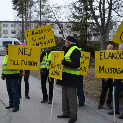 Nio personer står med gula plakat med sloganer mot en kommunsammanslagning.