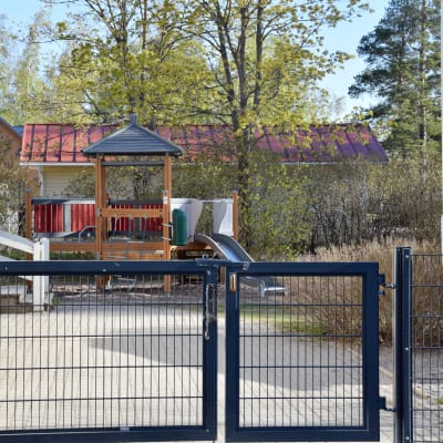 Gården på ett daghem. En stängd järngrind och inne på gården ser man en rutsckana och en klätterställning som liknar ett hus.