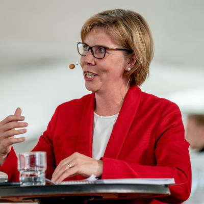 Anna-Maja Henriksson står vid ett bord och pratar under regeringens presskonferens i Helsingfors centrumbibliotek Ode den 3.6.2019.