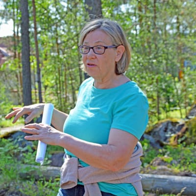 Kvinna står och förklarar något med händerna i en tallskog.