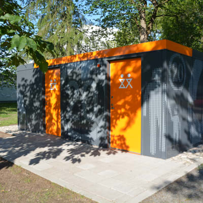 ny offentlig toalett i kuppisparken i åbo. grå rektangylär byggnad med orangea dörrar som stark kontrast.
