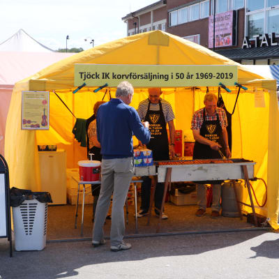 Tre personer står inne i ett gult tält och säljer korv.