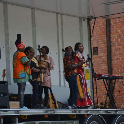 Fem kvinnor i färgglada kläder sjunger på en scen.