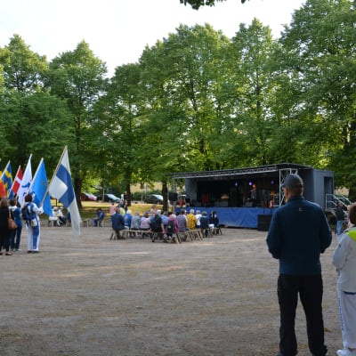 Personer står med flaggor vid en scen i en park.