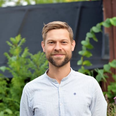 Jon Lindström, turismchef i Hangö, står i en vit murarskjorta framför en röd stugvägg med gröna växter.
