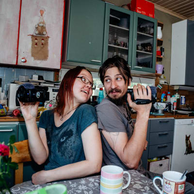 Hippihenkinen pariskunta istuu sotkuisessa ja värikkäässä keittiössä selät vastakkain videokamerat kädessä.