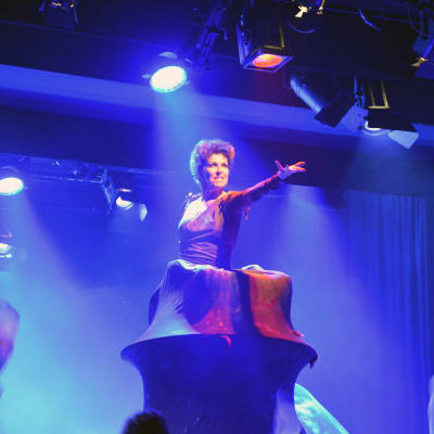 En kvinna på scen som står inne i ett "berg" med den enda armen utsträckt mot publiken.